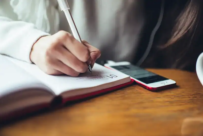 Person writing in planner with smartphone nearby.